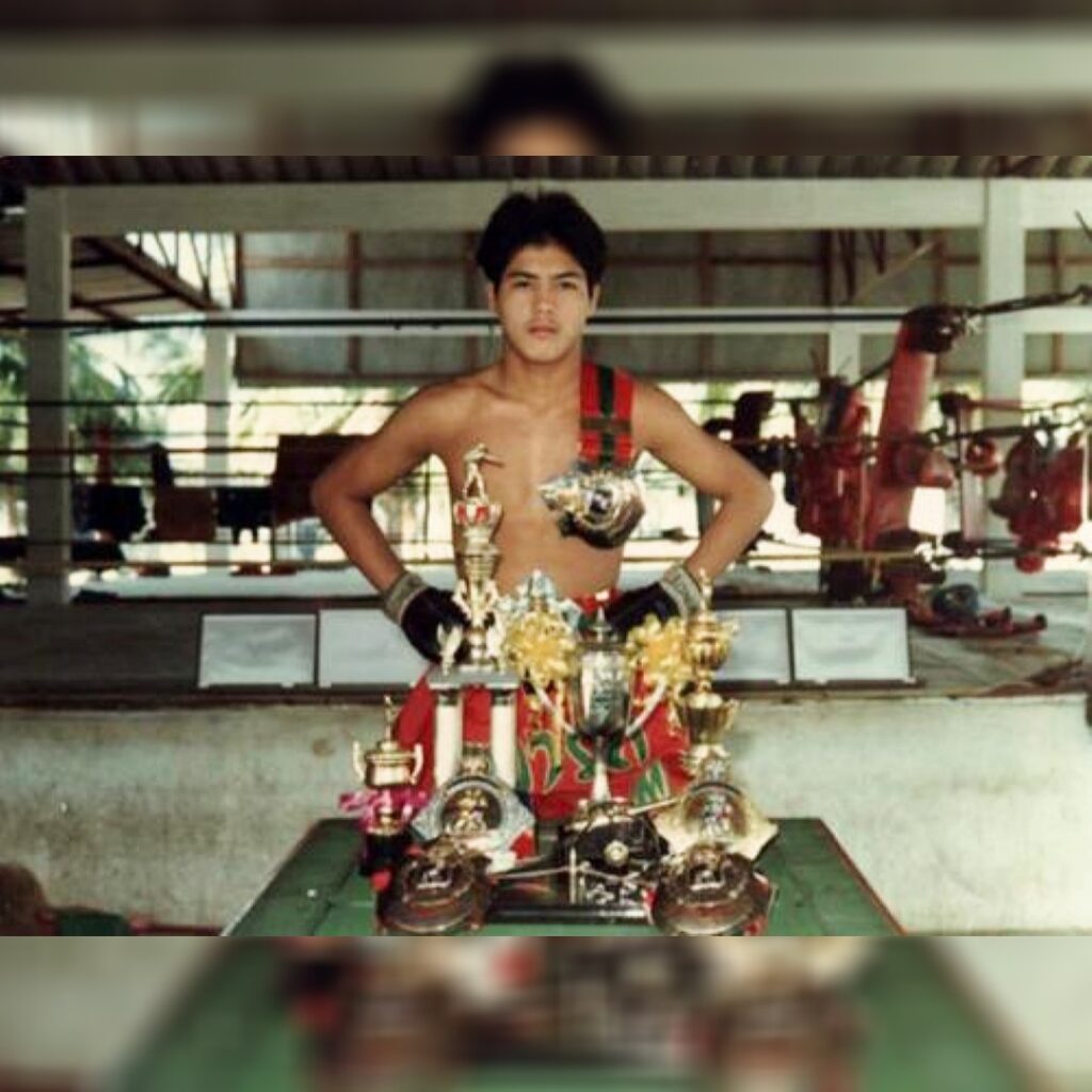 Samart Payakaroon standing with three Lumpinee belts and various other trophies